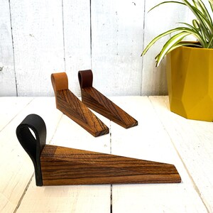 May include: Three wooden doorstops with leather loops. The doorstops are made of dark wood with a striped pattern. The loops are brown, black, and tan.