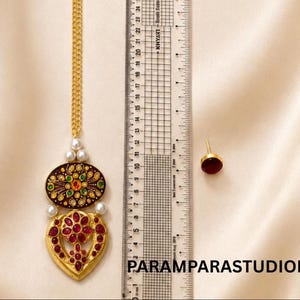 Puede incluir: Un maang tikka dorado con cadena, que presenta un colgante ovalado oscuro con detalles florales verdes y amarillos, una base en forma de corazón con piedras rojas y perlas blancas. Un pendiente de botón rojo se muestra junto a una regla de 24 cm.