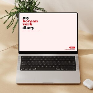May include: A laptop showing "my korean verb diary" in red and black text. The laptop is open on a light wooden surface, with a small plant and a framed picture in the background. White wireless earbuds and a black smartphone are also visible.