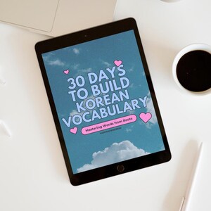 May include: A black tablet displays the text "30 DAYS TO BUILD KOREAN VOCABULARY" against a sky background with clouds and pink hearts. A laptop, a cup of coffee, a pen, and a decorative object are also visible on a white surface.