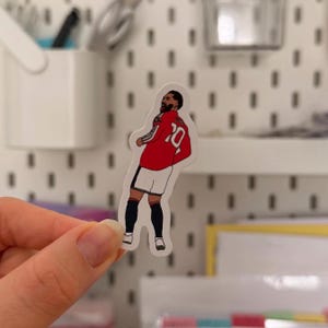 May include: A die-cut vinyl sticker featuring a cartoon illustration of a soccer player with a beard, wearing a red long-sleeve jersey with the number 10 on the back, white shorts, and dark socks, captured in a celebratory pose while looking sideways.