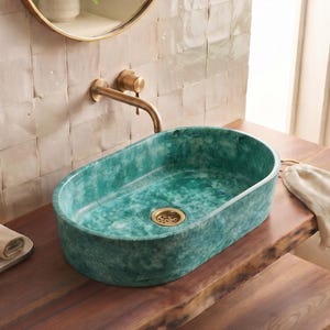 Peut inclure: Une vasque ovale en céramique de couleur sarcelle avec une finition marbrée et une bonde dorée, installée sur un comptoir de vanité en bois foncé avec un robinet mural assorti de couleur dorée au-dessus du lavabo.
