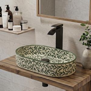 Puede incluir: Un lavabo ovalado de cerámica con un patrón de hojas verdes sobre fondo crema, montado sobre una encimera de madera con un grifo negro mate. Un estante flotante sostiene varios frascos de aseo y un espejo con marco de madera cuelga arriba.