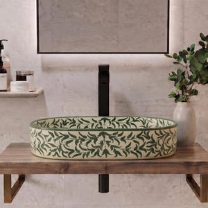May include: An oval vessel sink featuring a cream-colored base with a repeating dark green leaf pattern. The sink is mounted on a thick, natural-toned wooden shelf supported by two metallic brackets, paired with a tall, matte black vertical faucet.