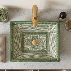 May include: A rectangular vessel sink featuring a green and white geometric pattern with a gold-toned tap. The sink is set into a speckled countertop alongside a small potted succulent, a beige textured towel, and a bar of soap on a dish.