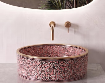 May include: A round ceramic vessel sink featuring a detailed pink and black floral pattern with a gold-toned rim. The sink is paired with a wall-mounted gold-toned faucet and handle, positioned above a white vanity with a marble countertop.