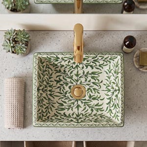 May include: A square ceramic vessel sink featuring a green leaf pattern on a cream background, paired with a gold tap. The sink is set into a speckled countertop alongside a potted succulent, a textured beige towel, and a bar of soap.