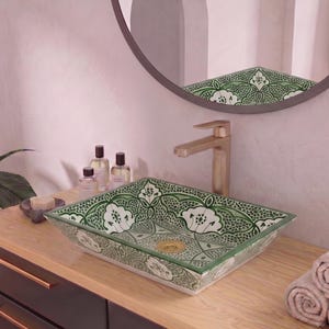 Puede incluir: Un lavabo rectangular de cerámica con un detallado patrón floral en verde y blanco. El lavabo cuenta con un grifo de oro cepillado y un desagüe a juego. Está montado sobre un mueble de madera con cajones oscuros y tiradores de color dorado.