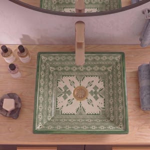 May include: A square ceramic bathroom sink featuring a cream-colored base with intricate green geometric and floral patterns. The sink includes a central gold-toned drain fixture and is paired with a tall, matching gold-toned faucet centered above.