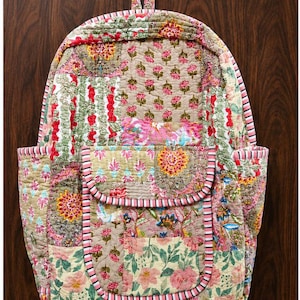 May include: A quilted backpack featuring a patchwork design of various floral and botanical patterns in shades of pink, green, yellow, and tan. The bag includes a front pocket with a flap, side pockets, and pink and white striped trim throughout.