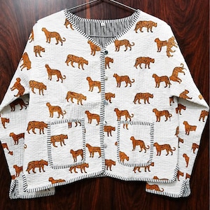 Könnte beinhalten: Eine weiße, gesteppte Jacke mit einem durchgehenden Druck von orangefarbenen und schwarzen Leoparden. Die Jacke hat einen V-Ausschnitt, lange Ärmel, zwei aufgesetzte Vordertaschen und eine Knopfleiste, alles mit schwarz-weiß gestreifter Einfassung.