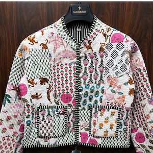 Könnte beinhalten: Eine gesteppte Jacke mit Patchwork-Design, verziert mit weißen, rosa, grünen und orangefarbenen Blumen- und Tigermotiven. Die Jacke hat einen Rundhalsausschnitt, lange Ärmel, zwei aufgesetzte Vordertaschen und eine Knopfleiste mit weißen Knöpfen.