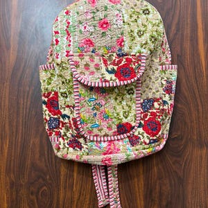Könnte beinhalten: Ein gesteppter Rucksack mit Patchwork-Design und Blumenmustern in Rot-, Rosa-, Grün- und Blautönen auf beigem Hintergrund. Die Tasche verfügt über eine Vordertasche mit Klappe, Seitentaschen sowie gestreifte Einfassungen an Kanten und Riemen.