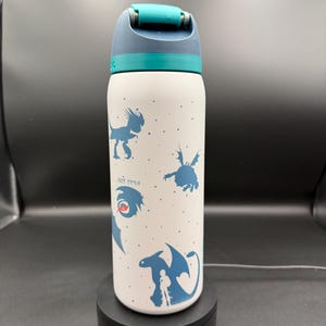 Puede incluir: Una botella de agua cilíndrica blanca con siluetas azules de varios dragones y un pequeño acento rojo en uno de ellos. La botella tiene una tapa abatible de color azul verdoso y azul oscuro. La superficie está decorada con puntos azules.