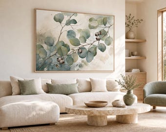 May include: A large rectangular wall art piece featuring a watercolour painting of eucalyptus branches with green leaves and small brown seed pods. The artwork is displayed in a light wood frame and hangs above a beige sectional sofa in a room.