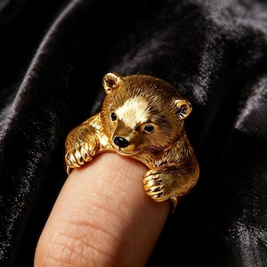 Könnte beinhalten: Ein goldfarbener Ring in Form eines Bärenjungen. Der Bärenkopf befindet sich oben, während die Vorderpfoten an den Seiten des Rings herabreichen. Der Bär weist eine detaillierte Fellstruktur, kleine schwarze Augen und eine schwarze Nase auf.