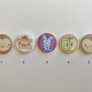 Pode incluir: Cinco botões redondos dispostos em uma linha. O primeiro e o quinto são bege com um rosto de desenho animado. O segundo mostra um bolo de morango. O terceiro é rosa com um coelho azul chorando e o texto ALWAYS CRYING. O quarto é uma maçã.