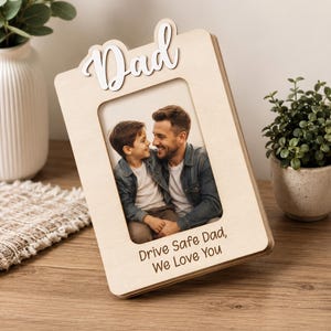 Puede incluir: Un marco de fotos de madera clara con la palabra Dad en letra cursiva blanca en la parte superior y el texto grabado Drive Safe Dad, We Love You en la parte inferior. El marco contiene una fotografía de un niño y un adulto sonriéndose.