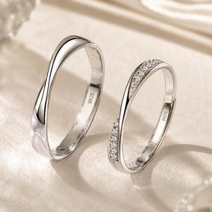 May include: Two silver-coloured rings with an S925 hallmark stamped on the inner band. One ring features a smooth, polished finish with a twisted design, while the other ring has a similar twisted shape accented with a row of small, clear stones.