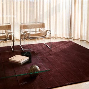 Op de afbeelding: Een bordeauxrood vloerkleed met een geometrisch patroon, een glazen salontafel en twee stoelen. Een boek en een decoratieve bol liggen op de tafel. De stoelen hebben een metalen frame en beige stof. Het kleed is ongeveer 2,5 x 3 meter.