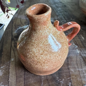 Op de afbeelding: Een kleine, gespikkelde keramische kruik met een afgeronde body en een handvat. De kruik is voornamelijk bruin met roodbruine spikkels en een rood-oranje handvat. De opening is breed en de kruik staat op een houten oppervlak. De stijl is rustiek en handgemaakt.