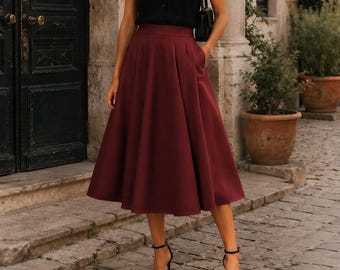 Puede incluir: Un top negro sin mangas con cuello en V y botones frontales, combinado con una falda midi plisada de cintura alta en color burdeos intenso. El conjunto se completa con un pequeño bolso de hombro negro y sandalias negras de tacón.