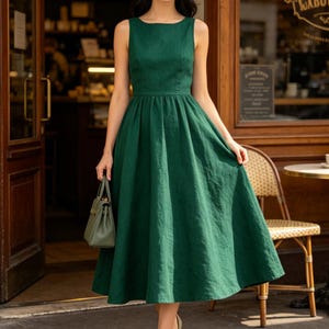 May include: A sleeveless forest green midi dress with a fitted bodice and a full, flared skirt. The dress features a round neckline and a textured fabric finish. A small, matching green structured handbag is held in one hand, and neutral-toned pointed-toe heels complete the ensemble.