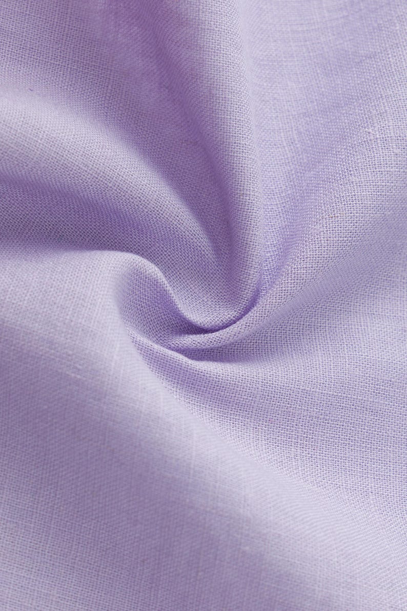 May include: A close-up view of light purple fabric with a visible woven texture. The material is gathered in the center, creating soft folds and shadows that highlight the consistent lavender hue and the natural weave of the textile fibers.