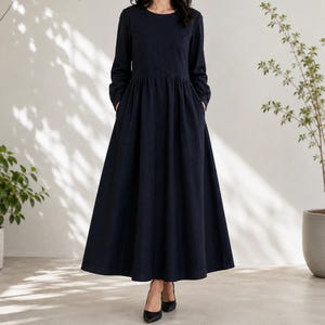 May include: A long-sleeved, floor-length dress in a solid dark navy blue color. The garment features a round neckline, a fitted bodice that transitions into a gathered waistline, and a full, flowing skirt that reaches the ankles with side pockets.