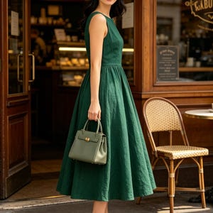 May include: A sleeveless dark green midi dress with a fitted bodice and a full, pleated skirt. The dress is paired with a small, structured sage green handbag featuring a gold-tone clasp and beige pointed-toe high-heeled pumps.