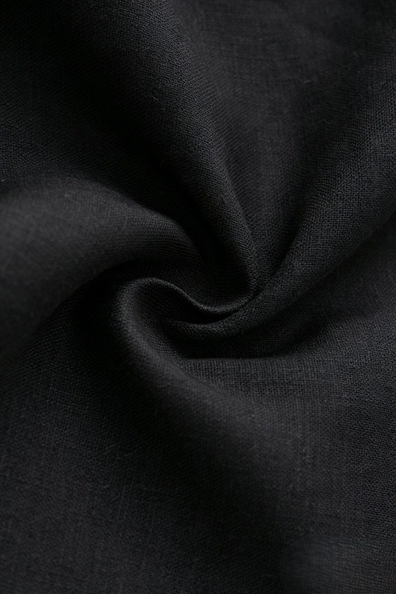 May include: A solid black fabric with a visible woven texture. The material is gathered into a central swirl, creating deep shadows and highlights that emphasize the weave and the matte finish of the dark textile surface in this view.