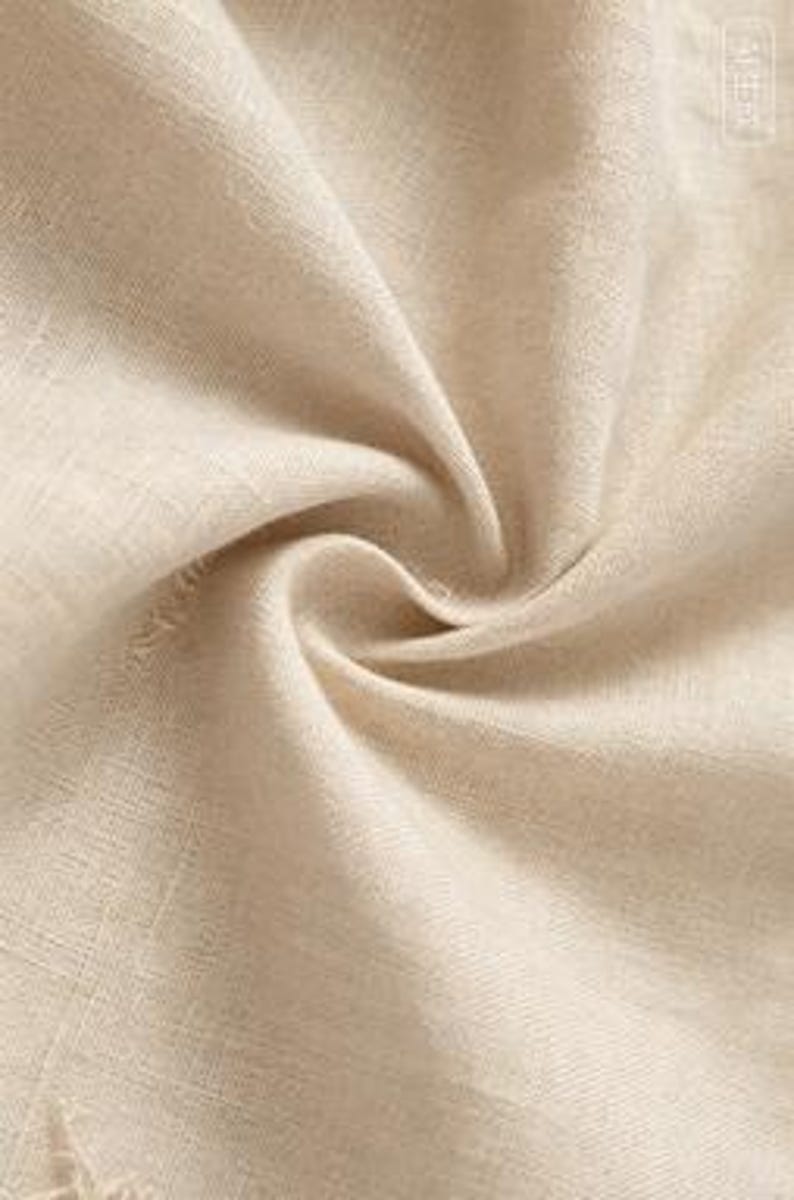 May include: A close-up view of beige linen fabric featuring a visible woven texture with natural fibers. The material is draped in soft, flowing folds, creating light and shadow across the surface. A small rectangular tag is visible in the corner.