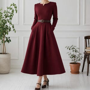 May include: A long-sleeved, ankle-length dress in a deep burgundy color. The dress features a fitted bodice with a notched neckline, a flared skirt, and a dark brown belt with a rectangular buckle cinched at the waist. The fabric appears smooth.
