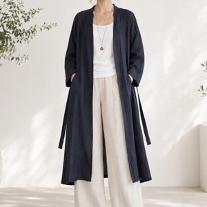 May include: A long, dark navy blue open-front duster coat with three-quarter length sleeves and side pockets, worn over a plain white sleeveless top and loose-fitting beige trousers, complemented by a simple gold pendant necklace on a thin chain.