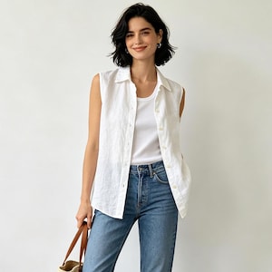 Può includere: Una camicia bianca senza maniche abbottonata indossata sopra una canotta bianca, abbinata a jeans blu. La camicia presenta un colletto classico e un'abbottonatura frontale completa. I jeans sono di un lavaggio medio con cinque tasche.
