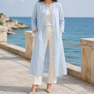 Può includere: Un cappotto lungo azzurro a maniche lunghe con scollo rotondo e chiusura frontale con bottoni. Il cappotto è indossato sopra un top bianco senza maniche e pantaloni bianchi, abbinato a décolleté beige con punta affusolata.