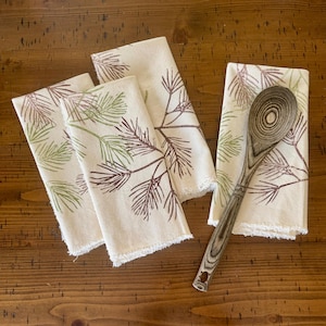 May include: Three off-white cloth napkins with frayed edges, each featuring a printed pattern of pine needle branches in muted green and brown tones, accompanied by a single wooden spoon with a visible dark wood grain pattern.