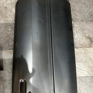Könnte beinhalten: Ein rechteckiges Autotürblech aus unlackiertem grauen Stahl. Das Blech hat eine glatte, flache Oberfläche mit einer vertikalen Vertiefung in der Mitte, einem rechteckigen Ausschnitt für den Türgriff und einem kleinen runden Loch unten.