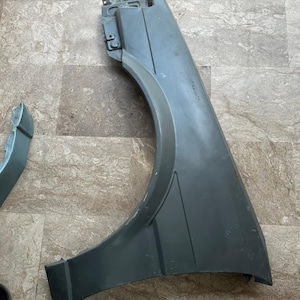 Peut inclure: Un panneau d'aile avant automobile gris présentant une découpe d'arche de roue incurvée, des supports de montage intégrés sur le dessus et une surface lisse et profilée. Une petite pièce de garniture grise détachée est située à gauche.