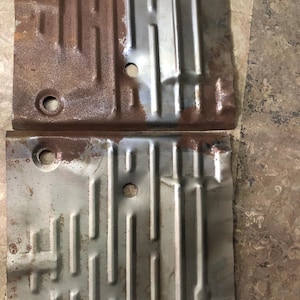 Könnte beinhalten: Zwei rechteckige Bodenbleche aus Metall mit geprägten Rippen zur strukturellen Verstärkung. Jedes Paneel zeigt eine Mischung aus oxidiertem braunem Rost und blankem silberfarbenem Stahl mit zwei kreisförmigen Löchern auf der linken Seite.