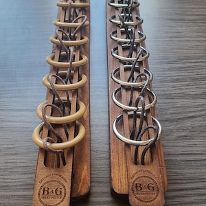 Könnte beinhalten: Zwei Holz-Denkspiele. Eines hat hellbraune Ringe und das andere silberne Ringe. Beide haben einen Holzrahmen mit einem Logo mit der Aufschrift "B&G Bead Puzzle". Die Puzzles befinden sich auf einer Holzoberfläche.