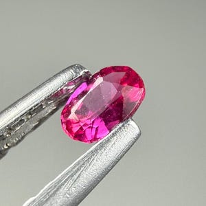 May include: A vibrant, oval-cut gemstone with a deep magenta-pink hue and multiple facets that reflect light. The stone is held by a pair of silver-toned metal tweezers, showcasing its translucent quality and polished surface in a clear, focused view.