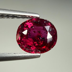 Op de afbeelding: Een gefacetteerde, ovaalvormige edelsteen in een diepe magenta en rode kleur, met talloze donkere interne insluitingen en spikkels door de steen heen. De edelsteen wordt vastgehouden door een zilverkleurige metalen pincet.