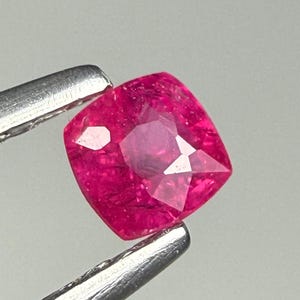 Op de afbeelding: Een levendige, kussengeslepen edelsteen met een diepe, verzadigde roze-rode tint. De steen heeft een gefacetteerd oppervlak dat licht reflecteert en de interne helderheid en structuur laat zien. Het wordt vastgehouden door een zilverkleurige metalen pincet.