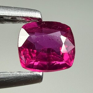 Op de afbeelding: Een edelsteen in kussenslijpvorm met een levendige, diepe magenta-roze tint en een gefacetteerd oppervlak dat licht reflecteert. De steen wordt vastgehouden door een metalen pincet, wat de transparante helderheid en de precieze geometrische snit toont.