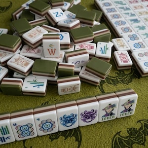 Puede incluir: Un conjunto de fichas de mahjong con caras blancas que presentan ilustraciones coloridas de bambú, caracteres y símbolos, junto con dorsos en capas de color verde, marrón y blanco. Algunas fichas muestran la palabra JOKER con una figura con sombrero cónico.