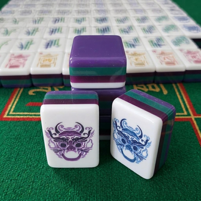 Może przedstawiać: Zestaw prostokątnych płytek do gry w Mahjong o warstwowej konstrukcji z jednolitym fioletowym wierzchem, zielonym środkowym paskiem i białą podstawą. Dwie płytki na pierwszym planie prezentują skomplikowane, stylizowane grawerunki gł&oacute;w smok&oacute;w w fioletowym i niebieskim tuszu.