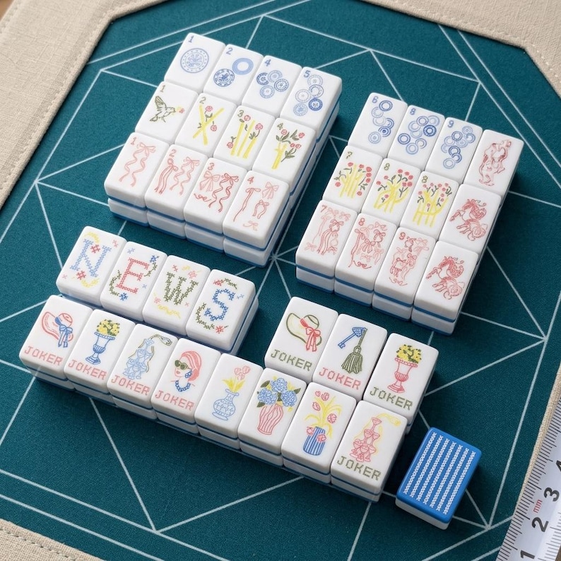 Op de afbeelding: Een verzameling witte rechthoekige Mahjong-tegels met kleurrijke kruissteekillustraties van bloemen, linten en het woord NEWS. Verschillende tegels zijn gelabeld als JOKER. Een blauwe tegel met witte strepen ligt bij een liniaal.