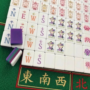 Może przedstawiać: Zestaw białych płytek do gry Mahjong ułożonych w rzędy, z kolorowymi literami kierunkowymi N, E, W, S, ilustracjami postaci Jokera oraz motywami kwiatowymi. Dwie luźne płytki leżą po lewej stronie, jedna w kolorze fioletowym i jedna w fioletowo-zielono-białe paski.