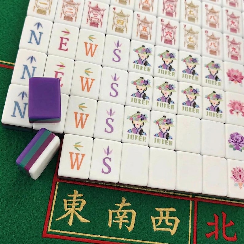 Może przedstawiać: Zestaw białych płytek do gry Mahjong ułożonych w rzędy, z kolorowymi literami kierunkowymi N, E, W, S, ilustracjami postaci Jokera oraz motywami kwiatowymi. Dwie luźne płytki leżą po lewej stronie, jedna w kolorze fioletowym i jedna w fioletowo-zielono-białe paski.